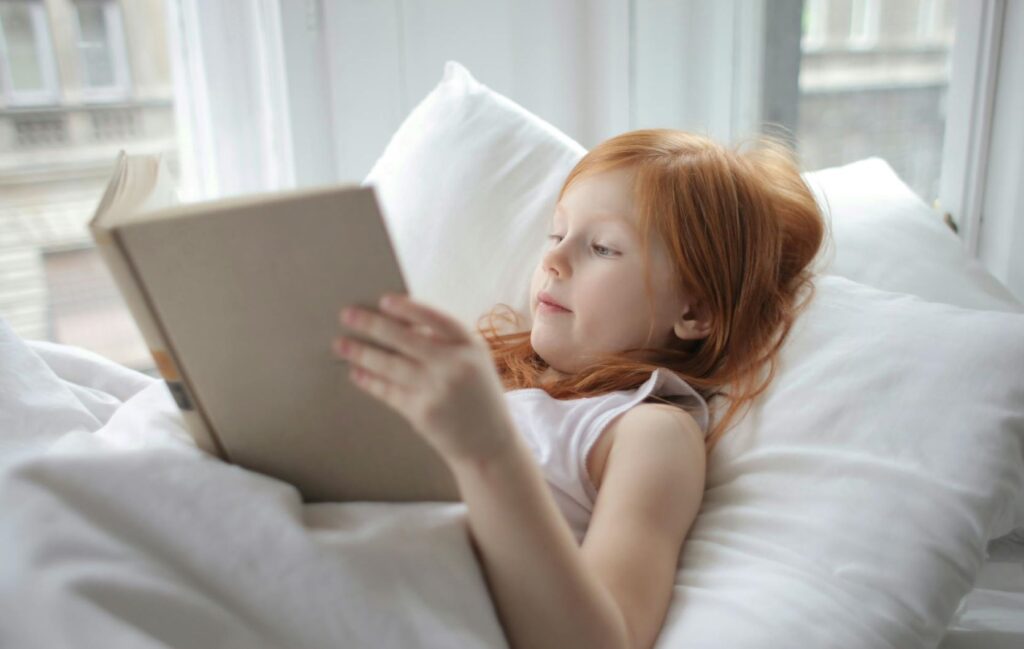 A young girl reading a book in her bed.